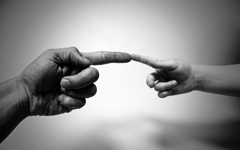Gros plan d'une main d'adulte et d'une main d'enfant qui se tendent et se touchent du bout des doigts sur un fond flou en noir et blanc, symbolisant la connexion - une image qui résonne avec ceux qui cherchent à comprendre les RH.