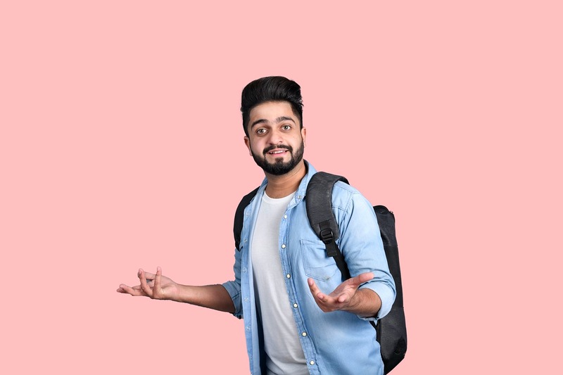 Un jeune homme barbu portant une chemise en jean et un sac à dos se tient devant un fond rose, faisant un geste des mains comme s'il demandait comment recruter un alternant en 2025.