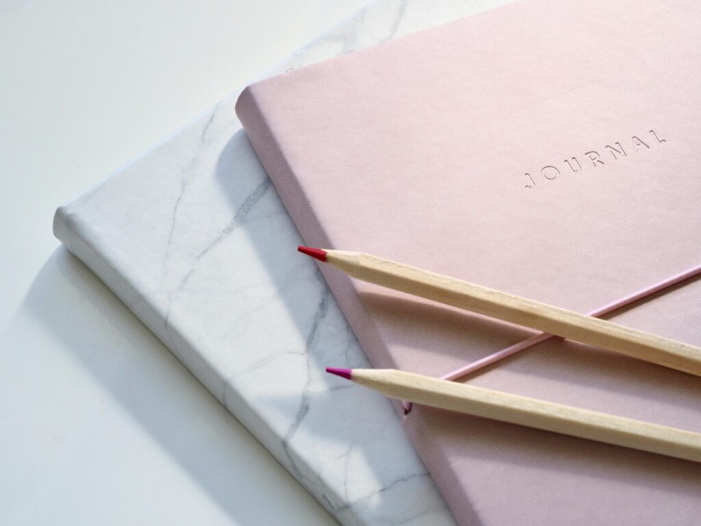 Deux crayons de couleur sont posés sur un journal rose avec la mention "JOURNAL" en relief sur la couverture, qui est empilé sur un cahier fermé à motifs de marbre blanc.