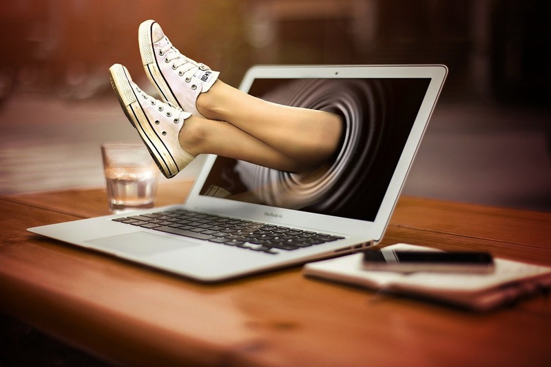 A pair of legs wearing white sneakers appear to emerge from the screen of a laptop open on a wooden table, symbolizing the innovative ways of recruiting a work-study student in 2025.