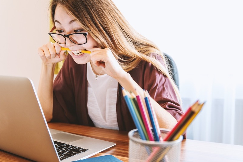 A woman sits at a desk, biting a pencil and staring frustratedly at her laptop screen, perhaps researching how to recruit a work-study student in 2025; a cup of colored pencils sits in the foreground.