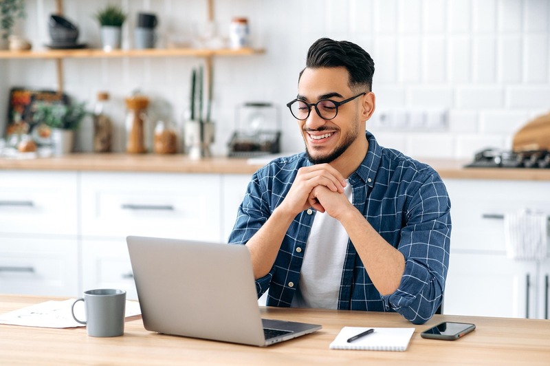 A man wearing glasses sits at a kitchen table, smiling at a laptop as he researches how to hire a work-study student in 2025. A laptop, phone, and mug are placed on the table. Shelves and kitchen utensils are in the background.
