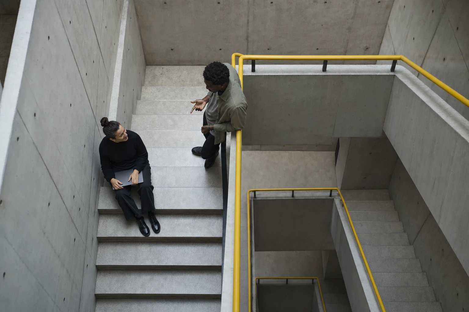 Looking down a stairwell: one person sitting on the steps, another standing at the railing